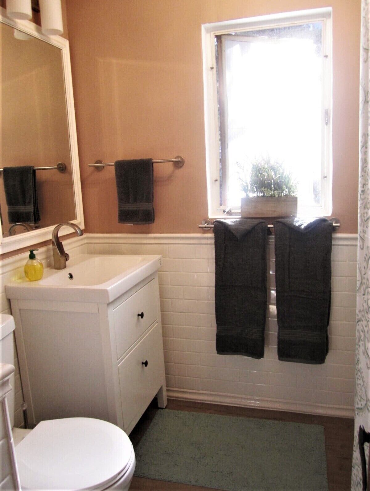 Ein Badezimmer mit einem weißen Waschbecken, einem Spiegel, einer Toilette und einem Fenster mit einem Topf auf der Fensterbank.