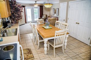 A dining area with a wooden table, white chairs, a chandelier, and a kitchen with a sink and cabinets.