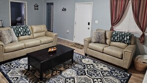 Una sala de estar con dos sofás beige, una mesa de centro negra, alfombras con diseños, una puerta blanca y una ventana con cortinas.