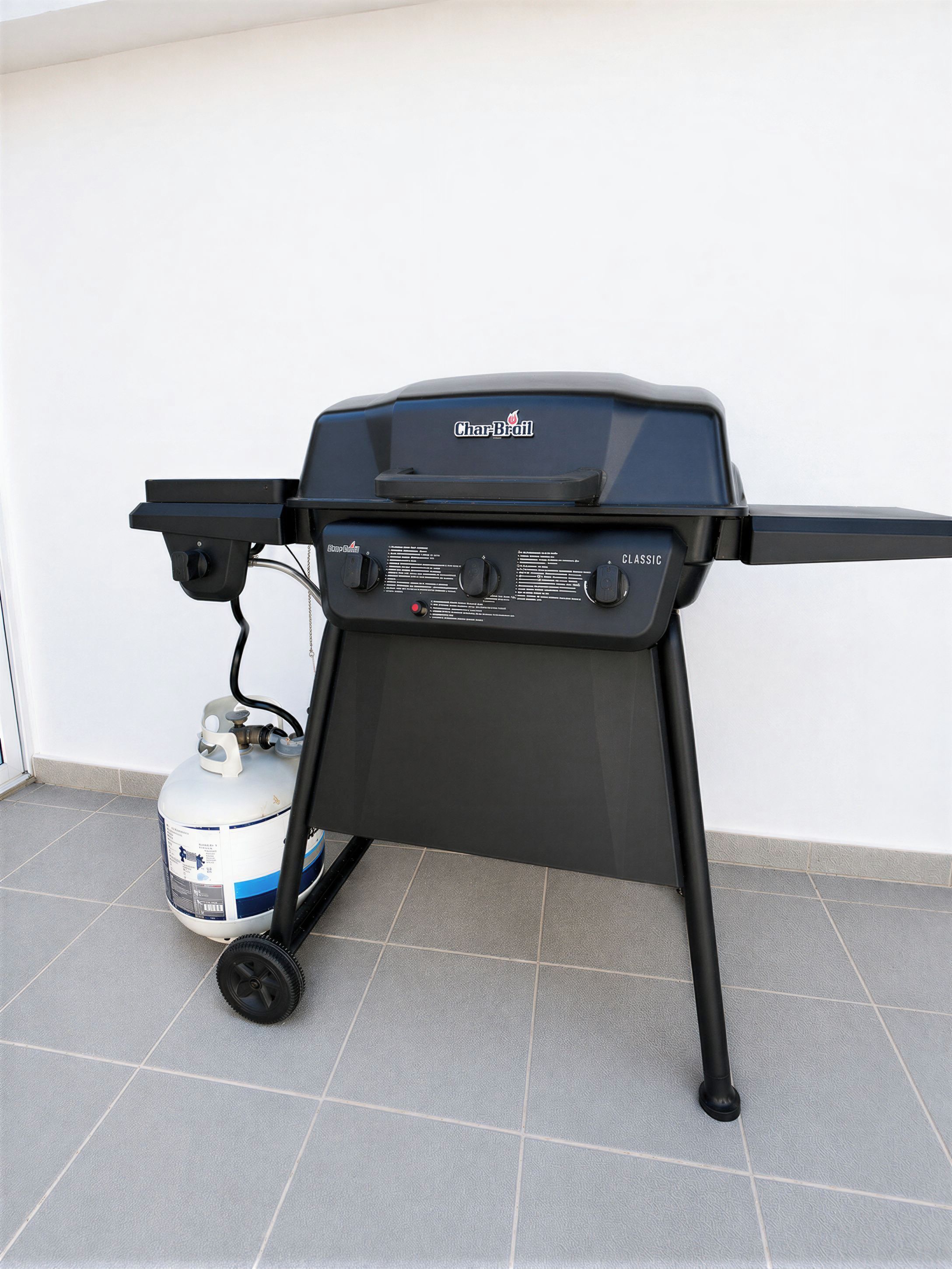 Ein Char-Broil Grill mit Propangasflasche, vor einer schlichten Wand auf Fliesenboden.