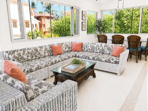 A spacious living area with a large sectional sofa, wicker furniture, and a view of a tropical landscape through large windows.