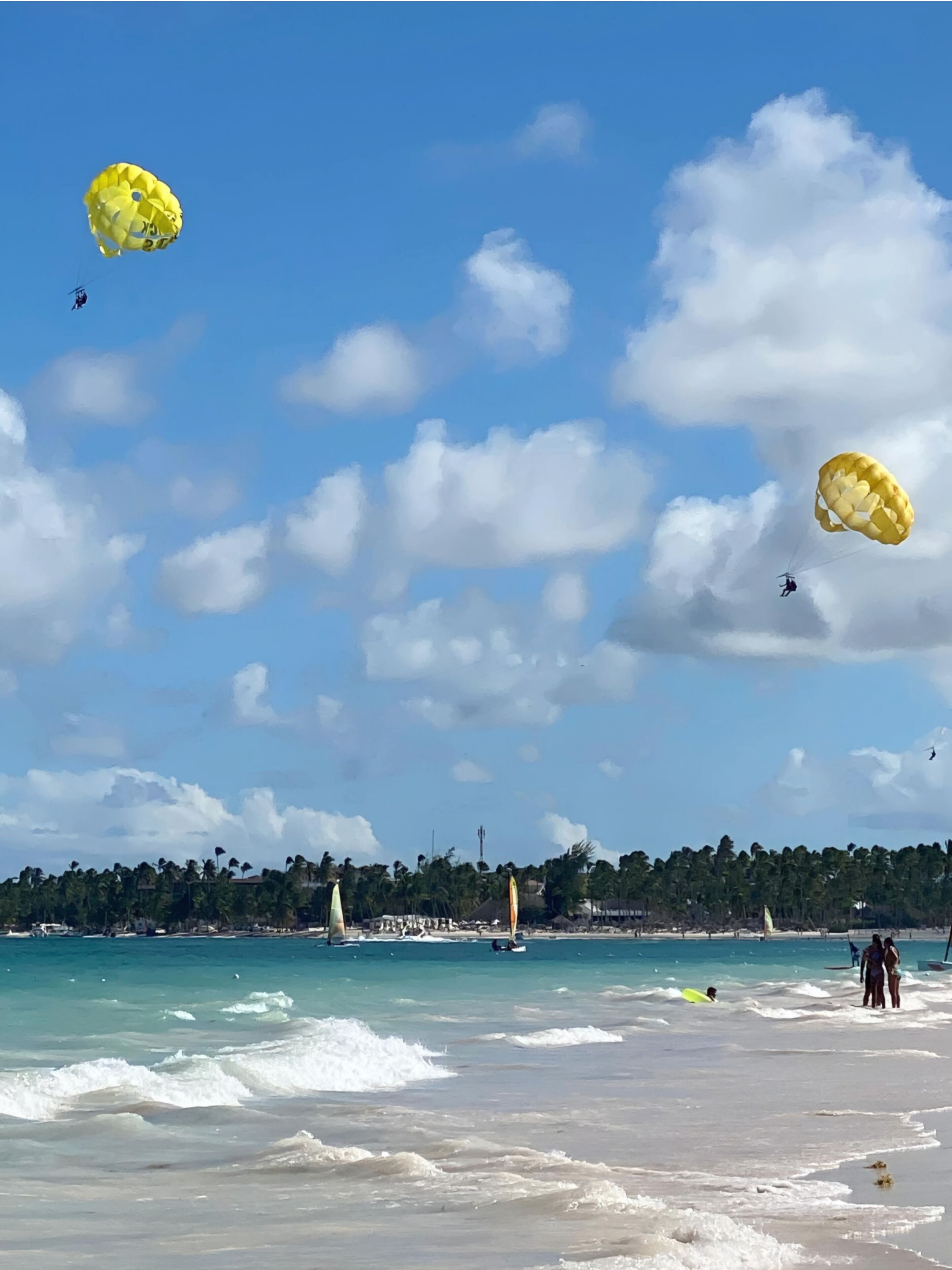 A beach with clear skies, two parachutes, and a distant shoreline with buildings and palm trees.