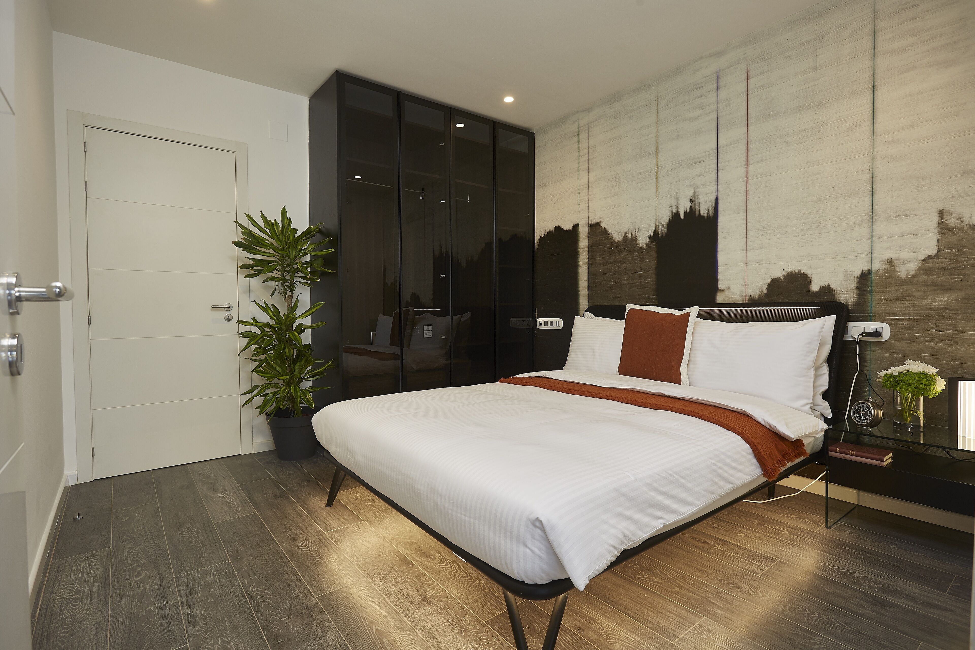 Une chambre moderne avec un grand lit, une plante en pot, une table de chevet et une fresque représentant un paysage urbain sur le mur.