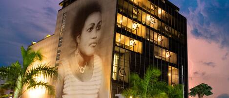 A building with a large mural of a person on its facade, palm trees in front, and a busy street with moving vehicles.