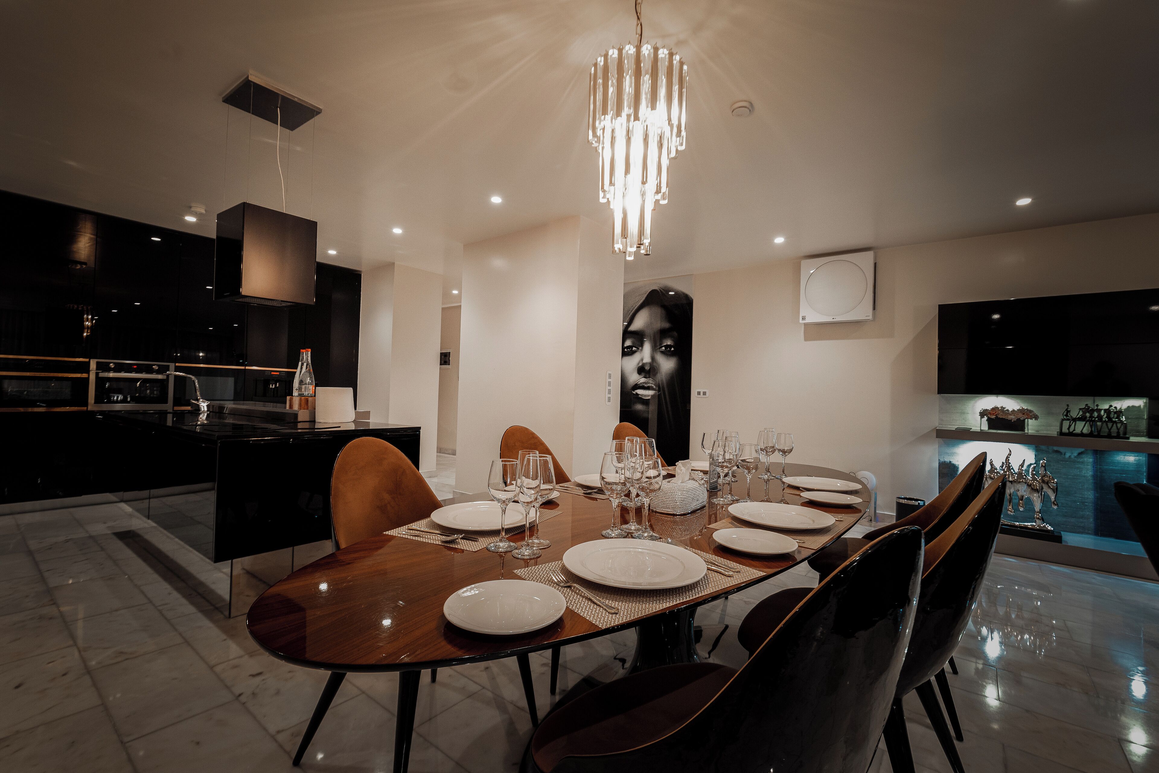 A modern dining area with a large wooden table set for six, surrounded by dark chairs. A chandelier hangs above, and a large portrait is on the wall.