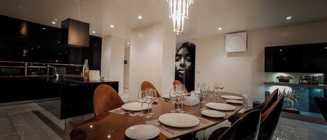 A modern dining area with a large wooden table set for six, surrounded by dark chairs. A chandelier hangs above, and a large portrait is on the wall.