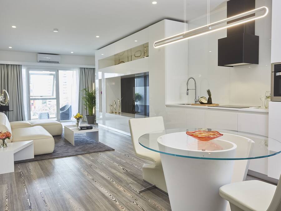 A modern living room with a glass-top dining table, a sofa, and a kitchen area with a built-in oven.