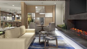 Ein modernes Wohnzimmer mit Sofa, Couchtisch und Sesseln. Ein Kamin mit Feuer, ein an der Wand befestigter Fernseher und eine Küchenzeile im Hintergrund sind zu sehen.