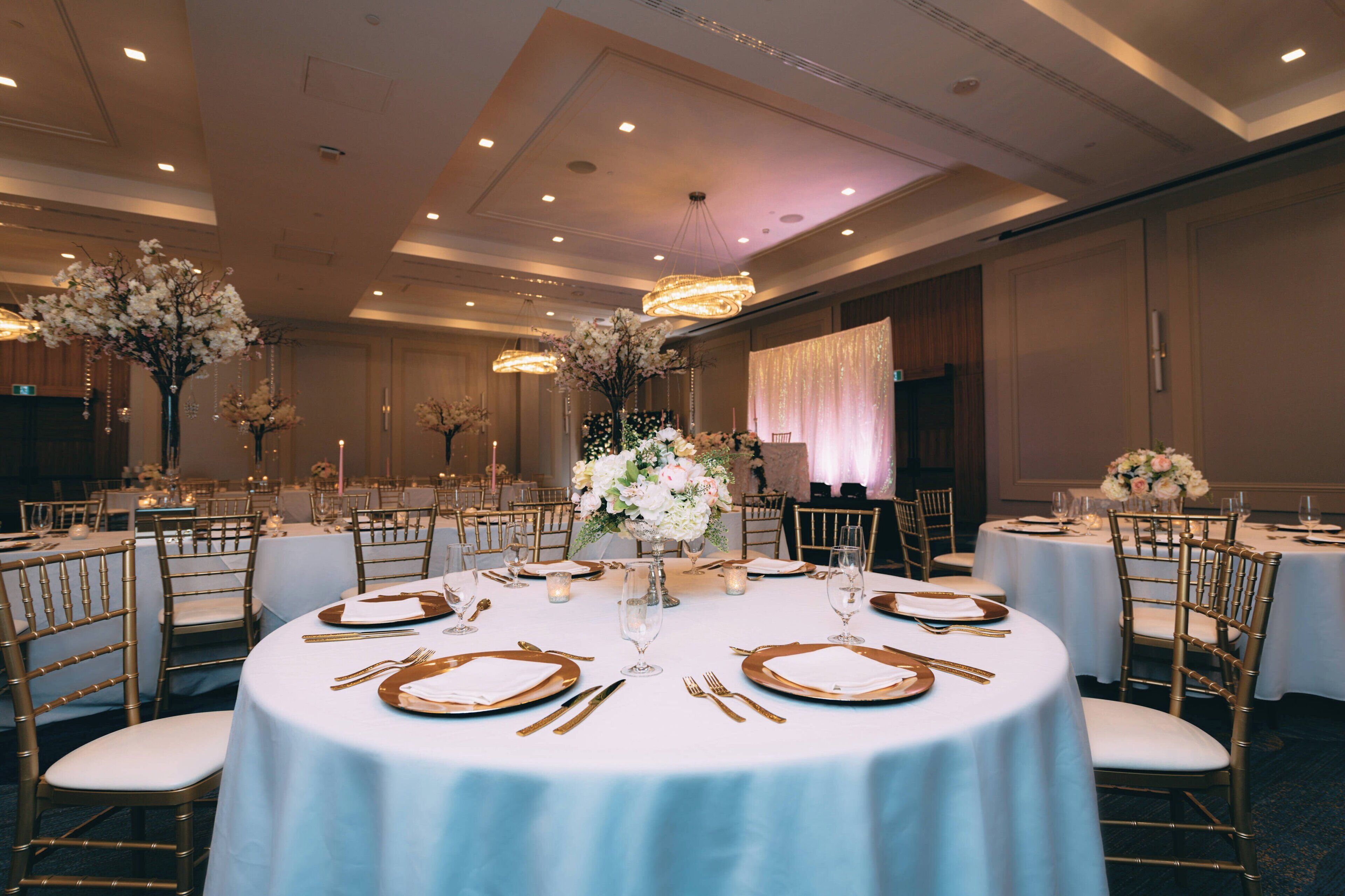 A banquet hall with round tables, gold chairs, white tablecloths, and floral centerpieces.