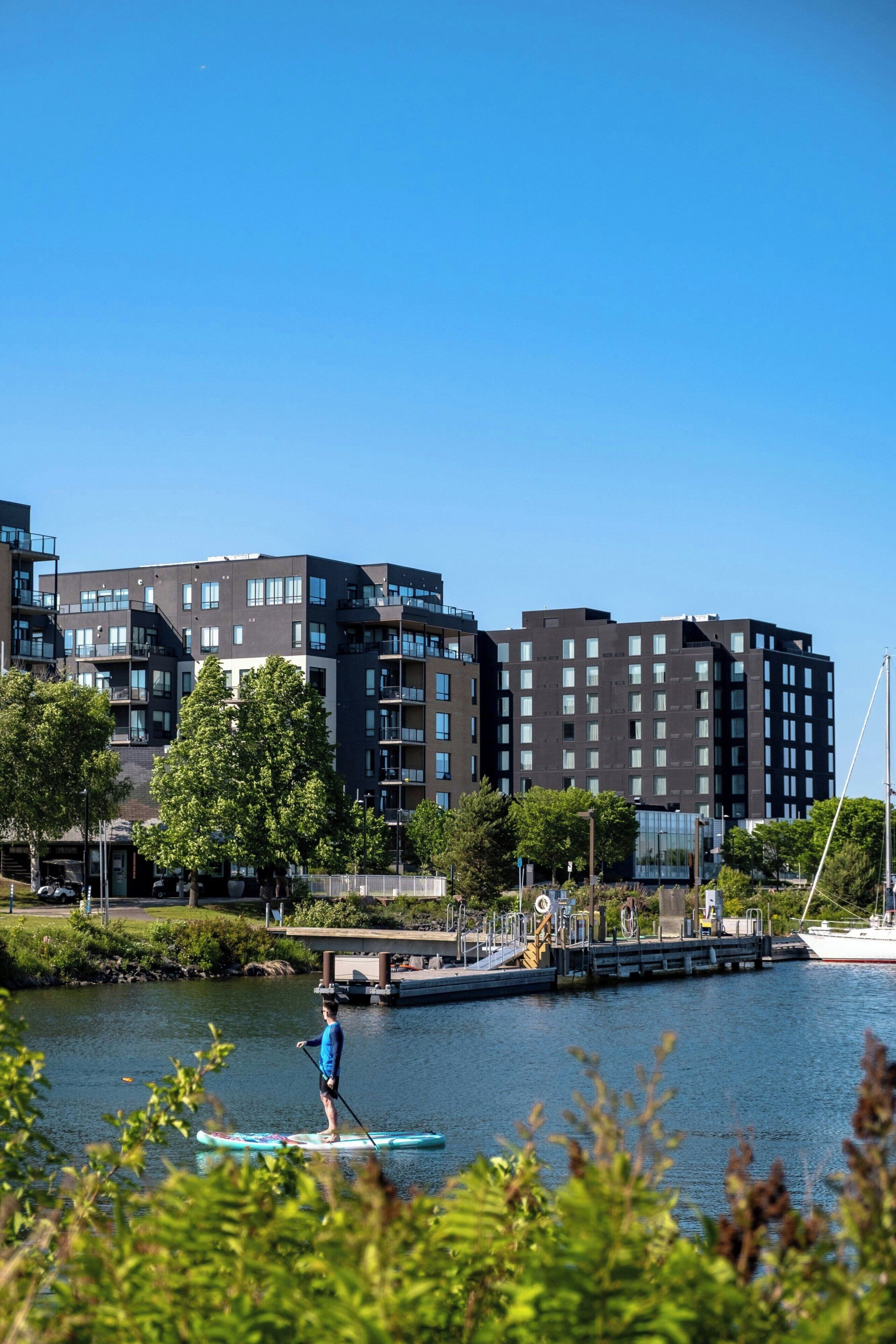 A modern building complex with multiple floors and balconies, a person paddleboarding on a calm body of water, and a dock with a boat.