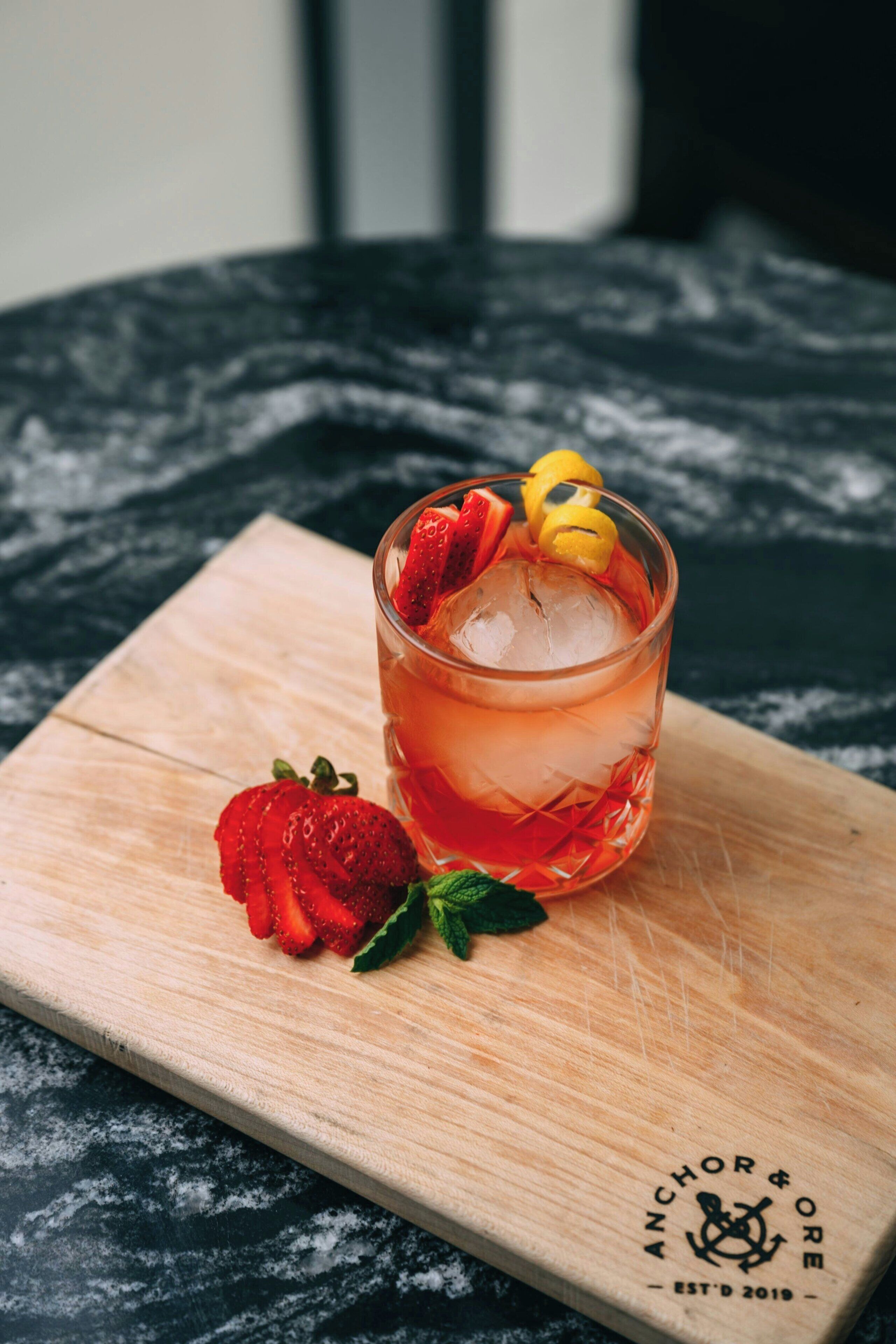 A glass of iced drink with strawberries and lemon slices on a wooden board.