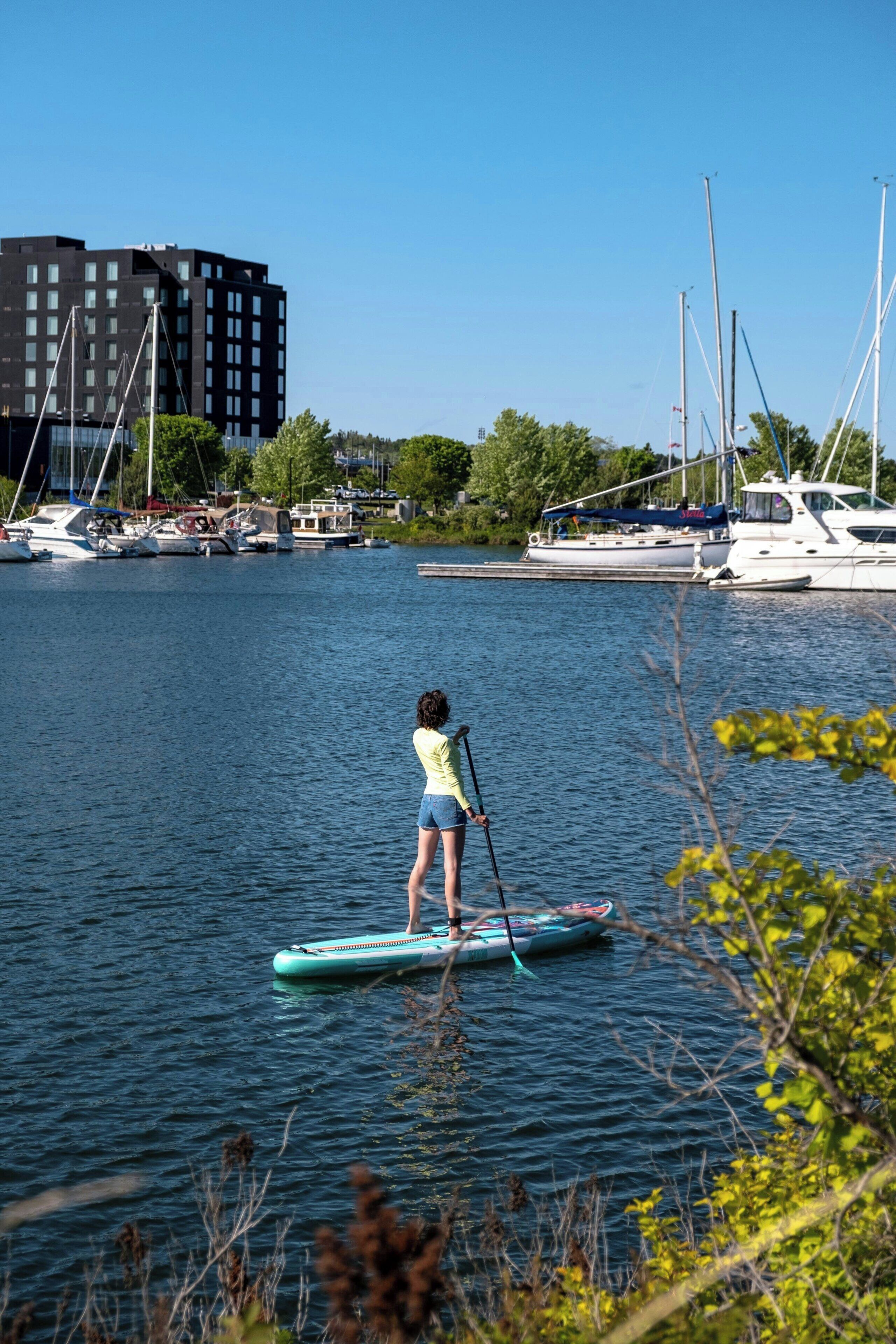 A person paddleboarding on a calm body of water with boats and a building in the background.