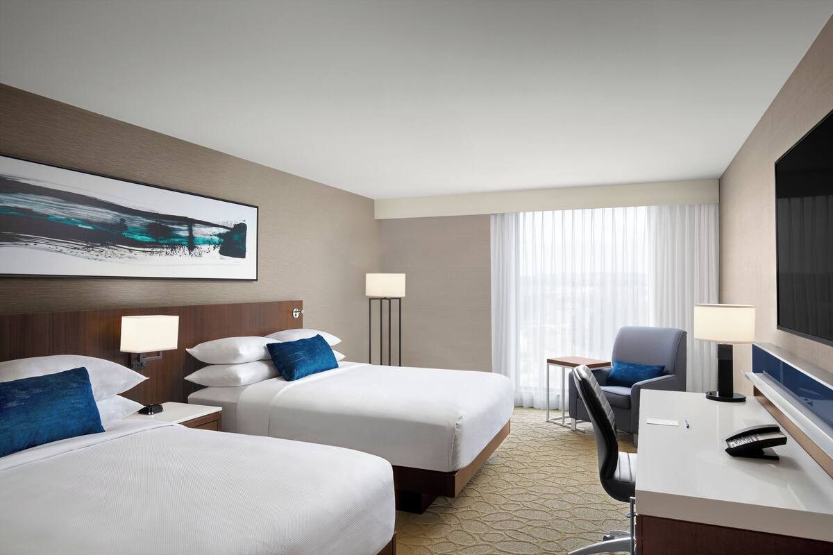 Une chambre d’hôtel avec deux lits, un bureau, une chaise, une télévision et une fenêtre avec des rideaux.