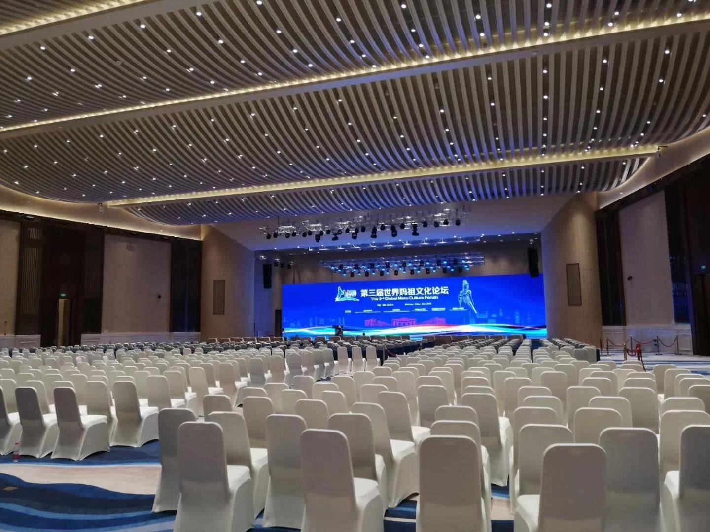 A conference room with rows of white chairs, a large screen at the front, and a modern ceiling design.