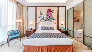 Habitación de hotel con una cama grande, una silla azul, un cuadro en la pared y un baño visible a través de una puerta de cristal.