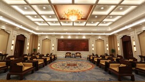 Un salón de conferencias con una gran mesa redonda, varias sillas, una alfombra estampada y un techo decorativo.