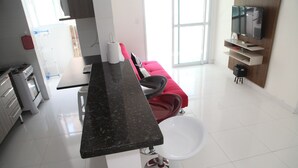 A modern kitchen with a black countertop, white sink, and red chair. There is a TV mounted on the wall and a window providing natural light. - 100m da Praia Aviação Imperdível (Praia Grande)