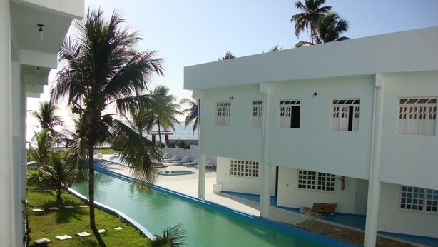 Un espace piscine agrémenté d’une fontaine aux formes arrondies, de palmiers et d’un bâtiment blanc doté de nombreuses fenêtres et de balcons.