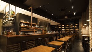 Un espace bar avec des tables et des chaises en bois, des étagères remplies de bouteilles et de verres, et un mur en briques.