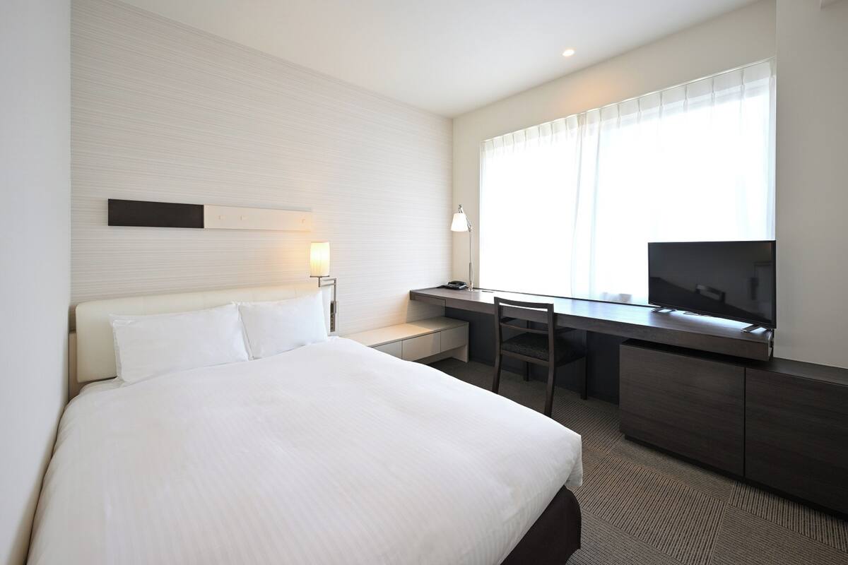 Una habitación de hotel con una cama grande, un escritorio con silla, televisor y ventana con cortinas.