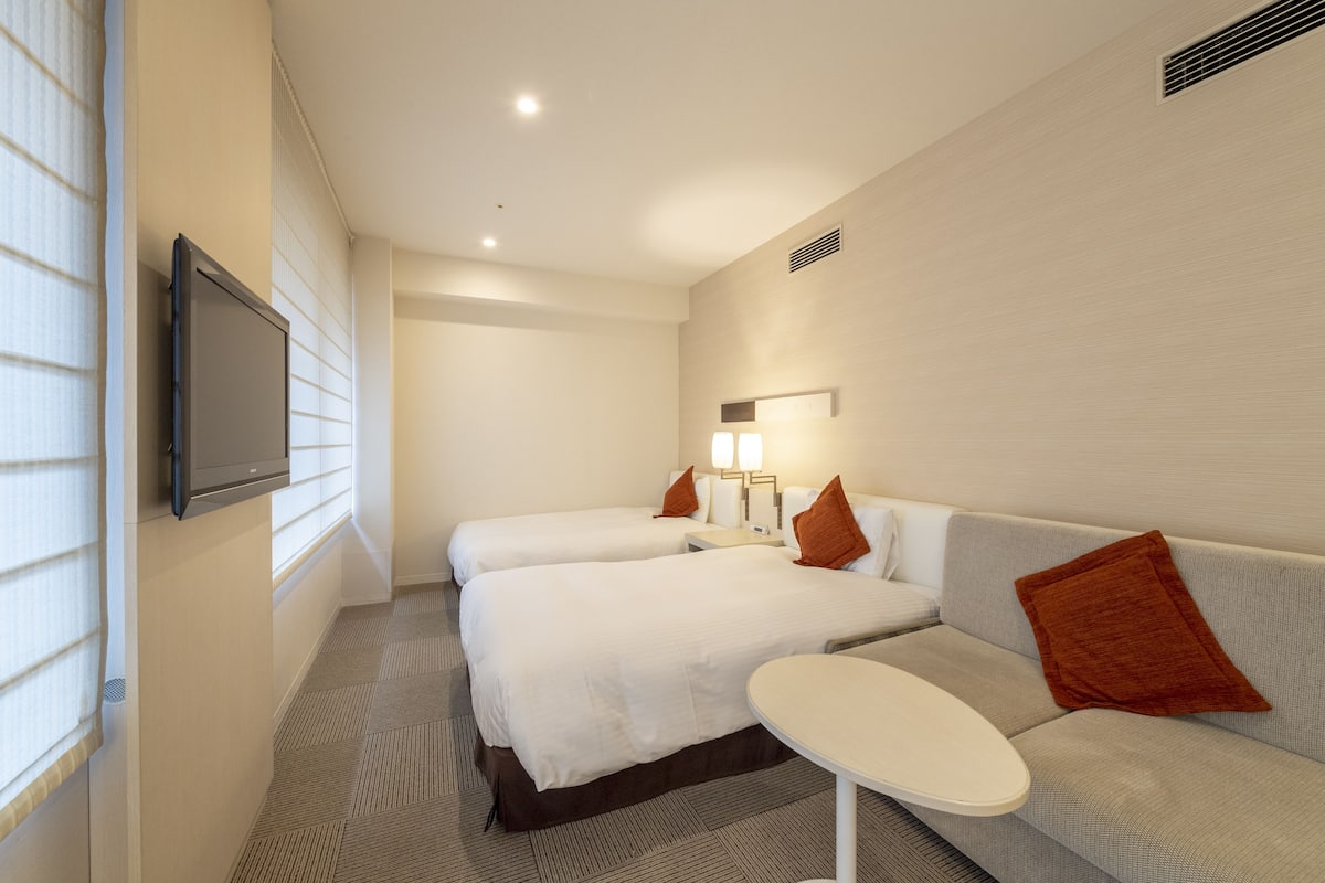 Una habitación de hotel con dos camas, un sofá, una mesita y un televisor montado en la pared.