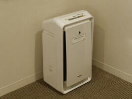 A white portable air purifier with a digital display, placed on a carpeted floor against a plain wall.
