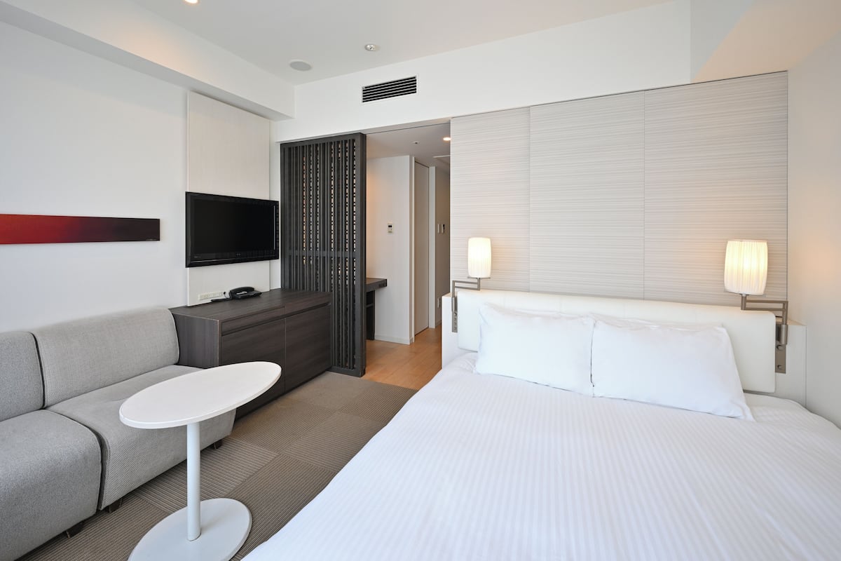 Una habitación de hotel moderna con una cama, un sofá, una mesita, un televisor y una lámpara.