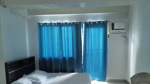 A bedroom with a bed, a chair, a window with blue curtains, and an air conditioning unit on the wall. - Angel Travellers Transit House (Parañaque)