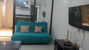 A teal sofa with two cushions, a flat-screen TV mounted on the wall, and a small table with a remote control and a decorative item. - Angel Travellers Transit House (Parañaque)