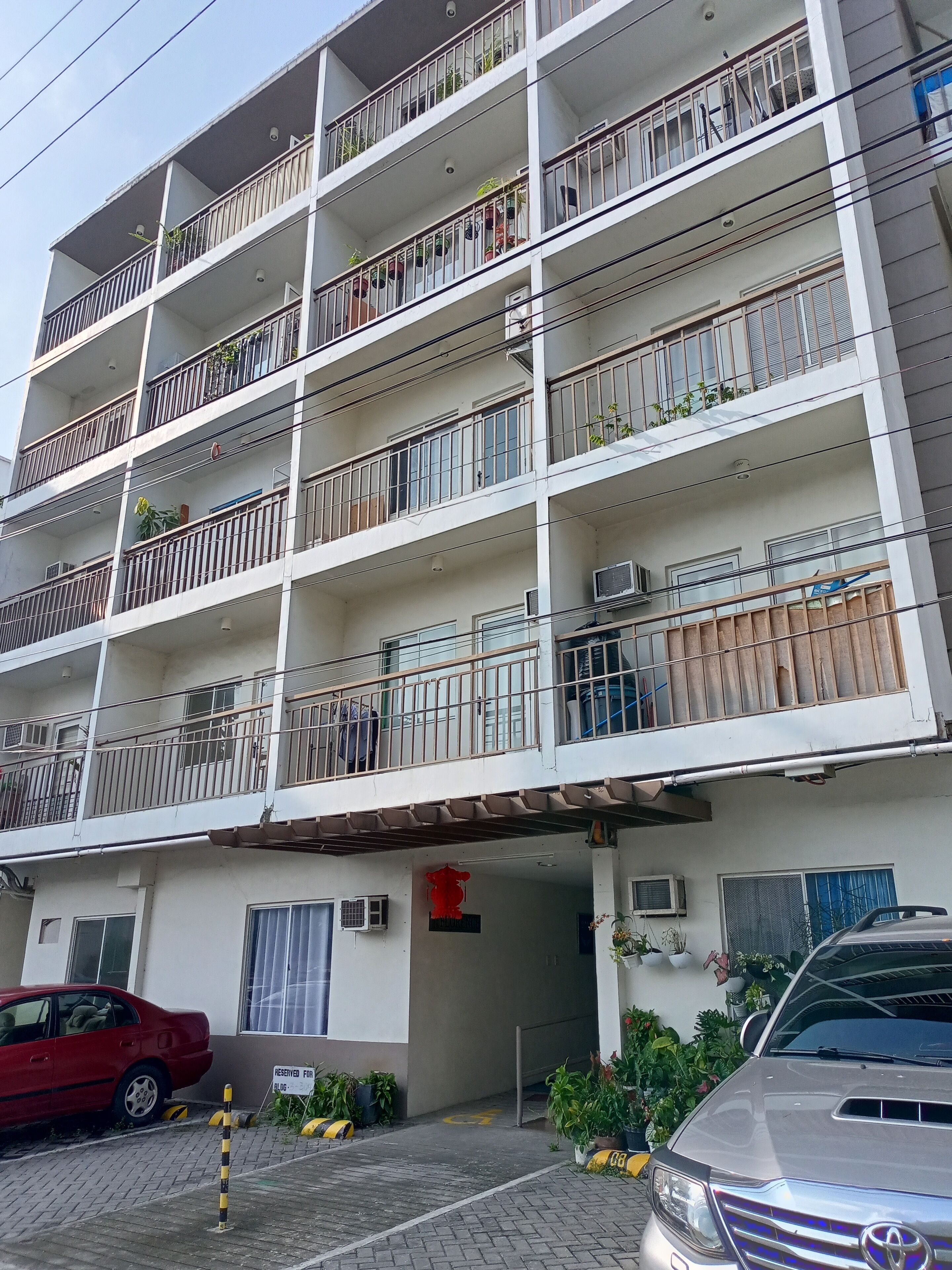 A multi-story residential building with balconies, air conditioning units, and a parking area with a red car and a silver SUV.