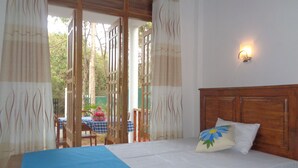A bed with a blue bedspread and a wooden headboard, a nightstand with a lamp, and a view of an outdoor area with a table and chairs. - Jungle View Guest (Polonnaruwa)
