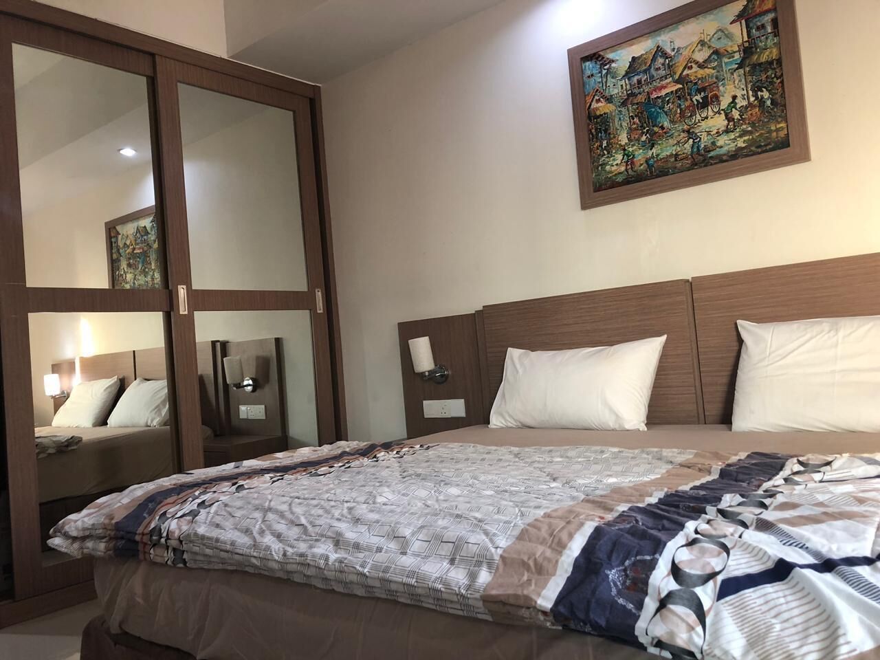 A hotel room with a large bed, a wardrobe with a mirror, a bedside table with a lamp, and a painting on the wall.