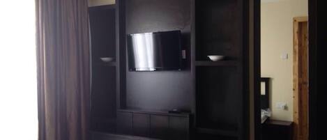Habitación de hotel con cama, televisor de pantalla plana montado en una pared de madera oscura, espejo y ventana con cortinas.