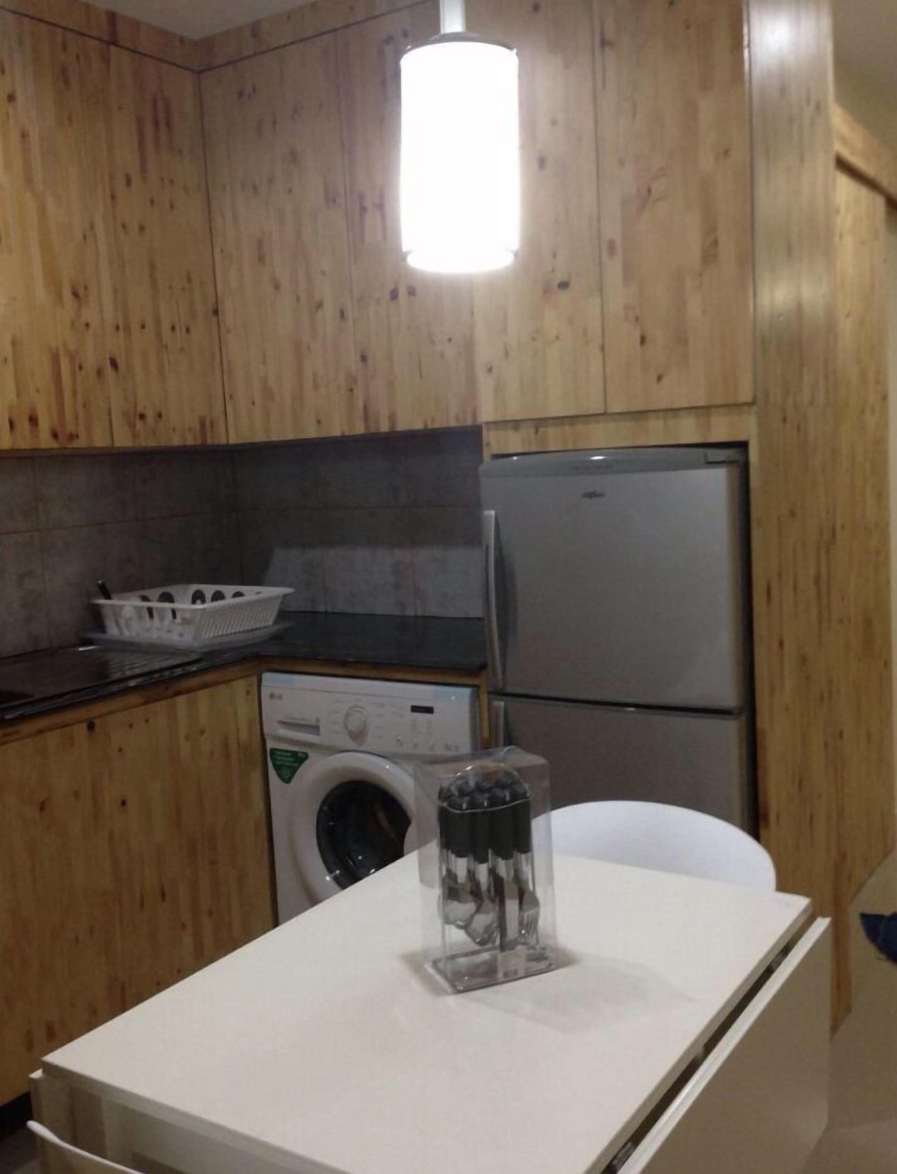 A compact kitchen with wooden cabinets, a washing machine, a refrigerator, and a small table with a glass container.