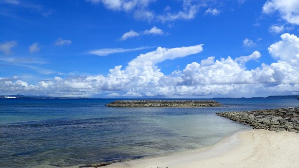 A beach with clear blue water, a rocky breakwater, and a sandy shore. - Smilax Kurichi (Uruma)