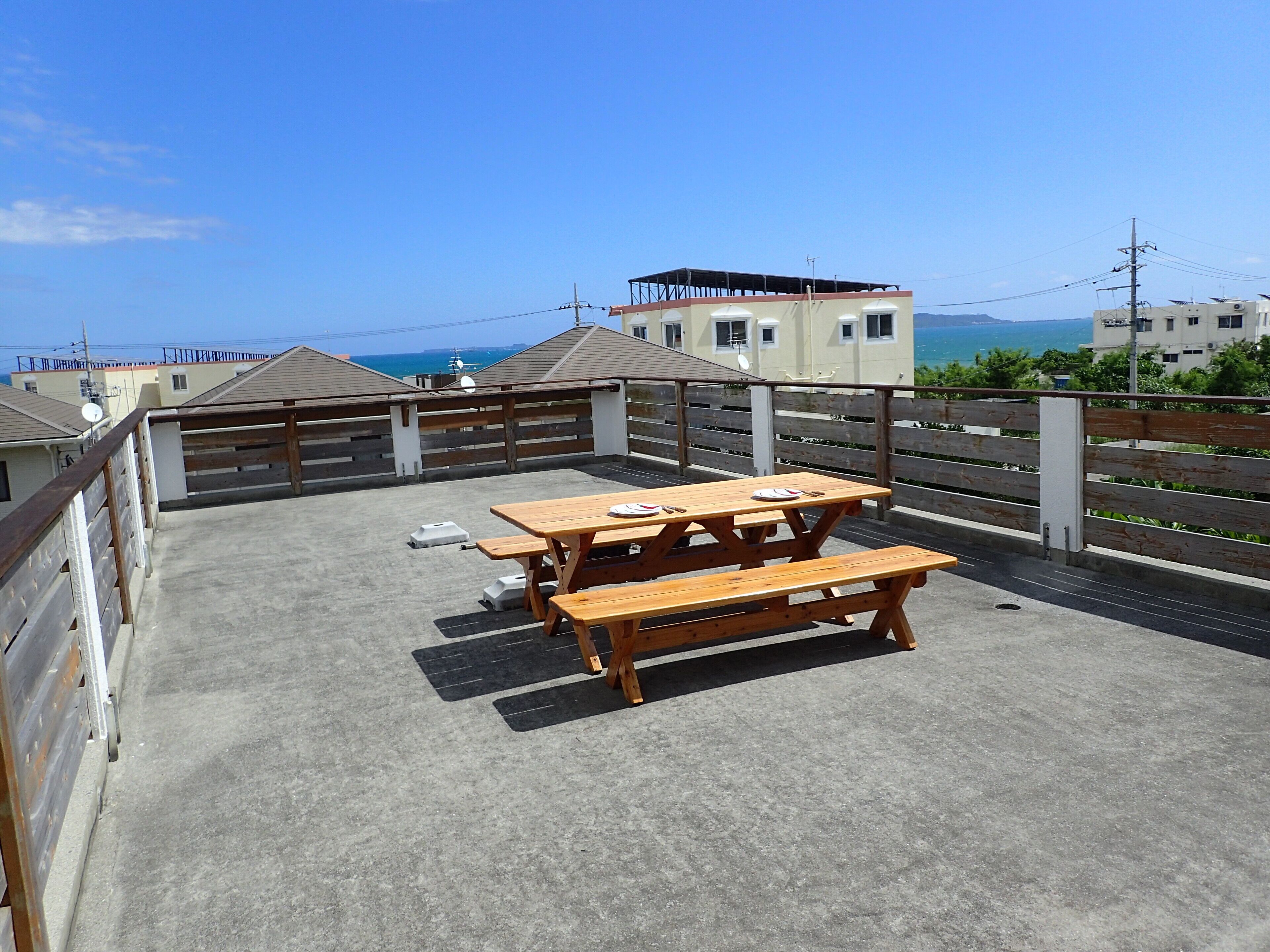 Eine Dachterrasse mit Holztisch und -bänken, einem Betonboden und Blick auf Gebäude sowie das Meer.