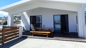 A rooftop terrace with a wooden table and benches, a glass door, and a white building with a chimney. - Smilax Kurichi (Uruma)