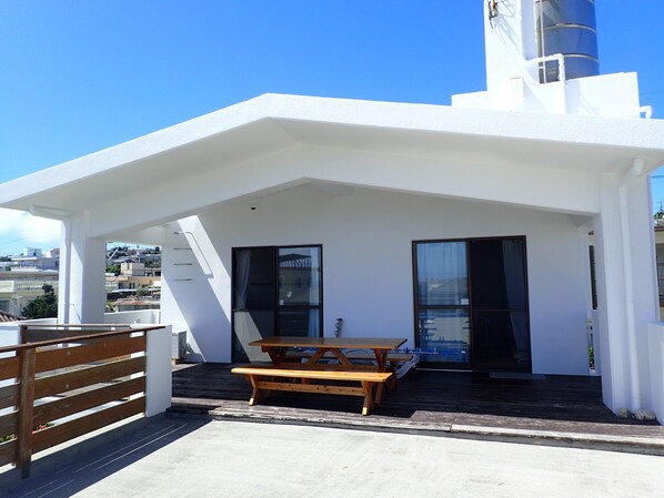 A rooftop terrace with a wooden table and benches, a glass door, and a white building with a chimney. - Smilax Kurichi (Uruma)