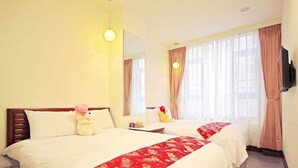Quarto de hotel com duas camas, mesa de cabeceira, luminária, televisão e janela com cortinas.