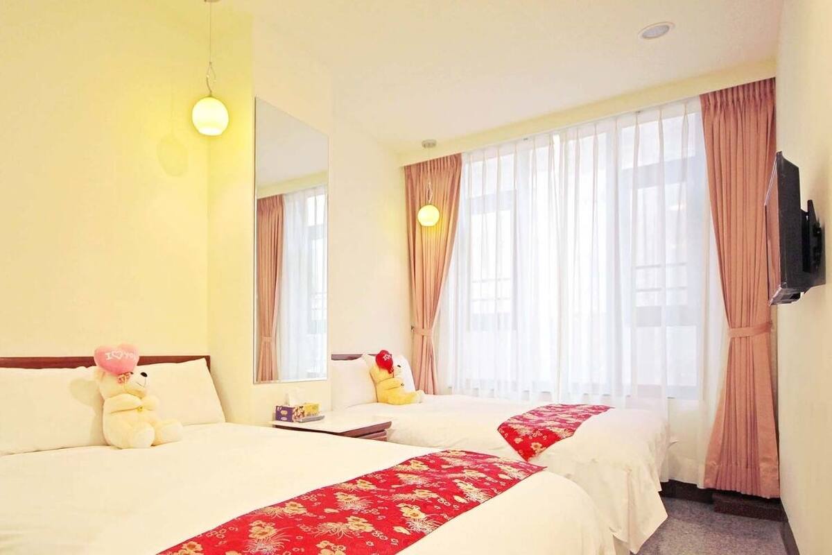 Ein Hotelzimmer mit zwei Betten, einem Nachttisch, einer Lampe, einem Fernseher und einem Fenster mit Vorhängen.
