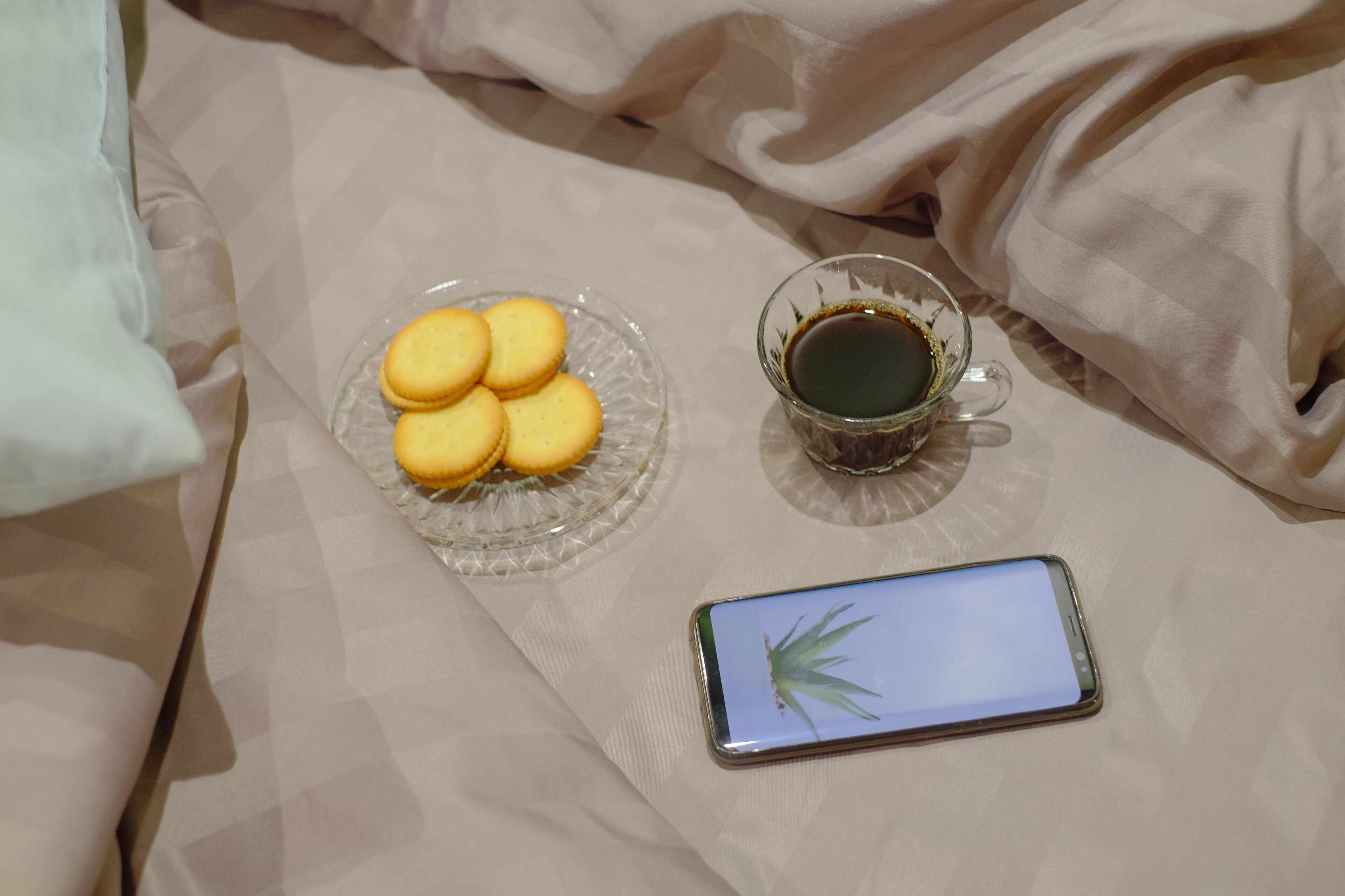 En seng med en glasskål fyldt med småkager og en kaffekop i glas, samt en smartphone på et hvidt lagen.