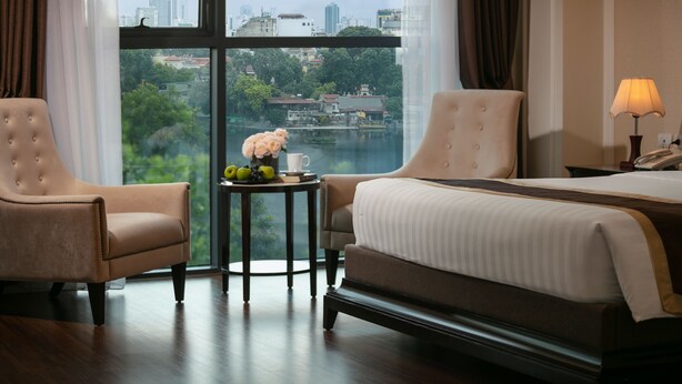 Ein Hotelzimmer mit großem Fenster, zwei Sesseln, einem kleinen Tisch mit Blumenvase und einem Bett mit Lampe.