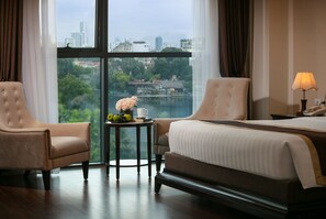 A hotel room with a large window, two armchairs, a small table with a vase of flowers, and a bed with a lamp.