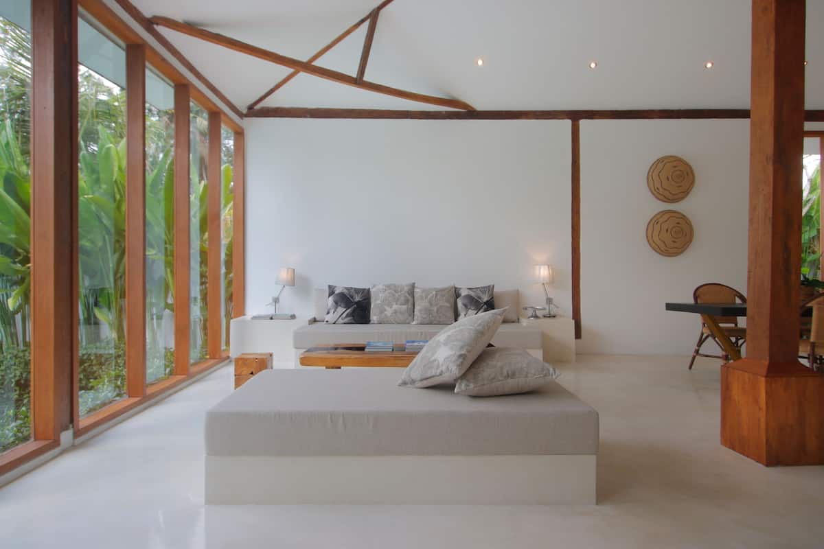 A modern bedroom with a large bed, a wooden ceiling, and large windows offering a view of greenery.