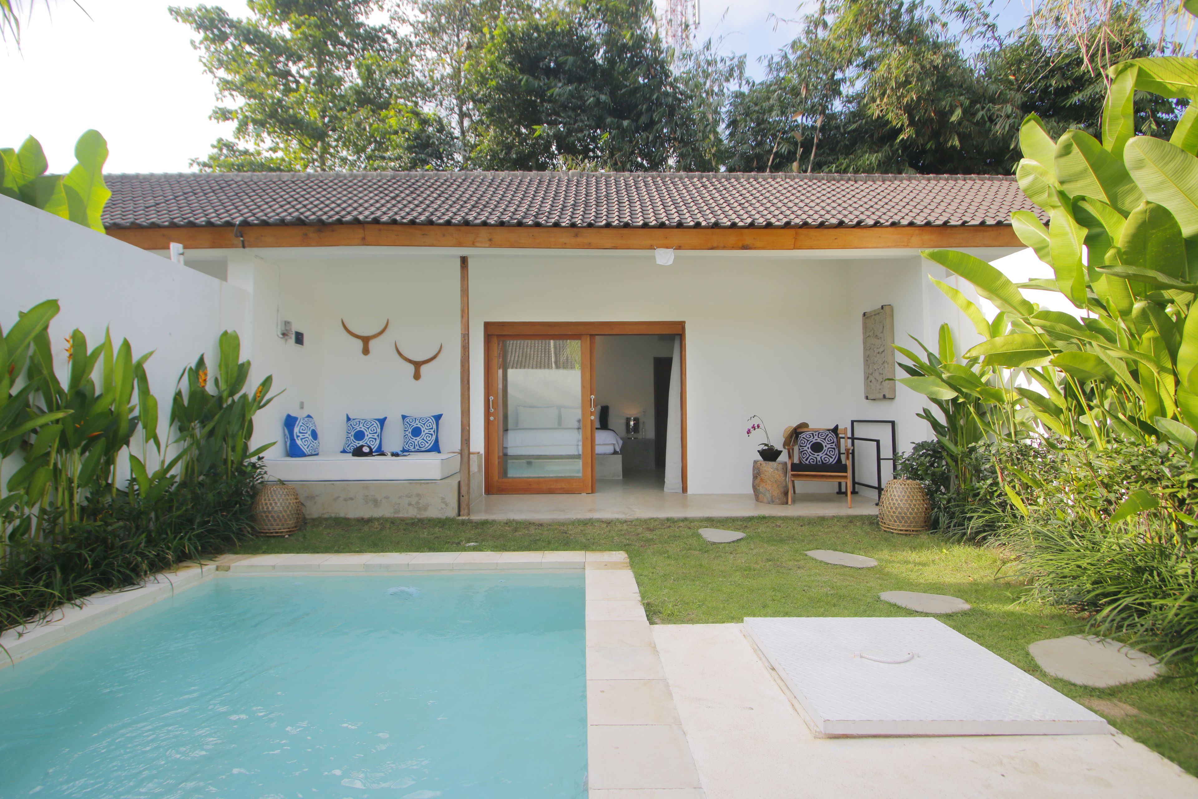 A poolside area with a clear blue pool, a small garden with stepping stones, and a building with a tiled roof and a door leading to a room.