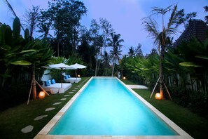 Una piscina rectangular con superficie de agua azul cristalina, rodeada de exuberante vegetación y palmeras.
