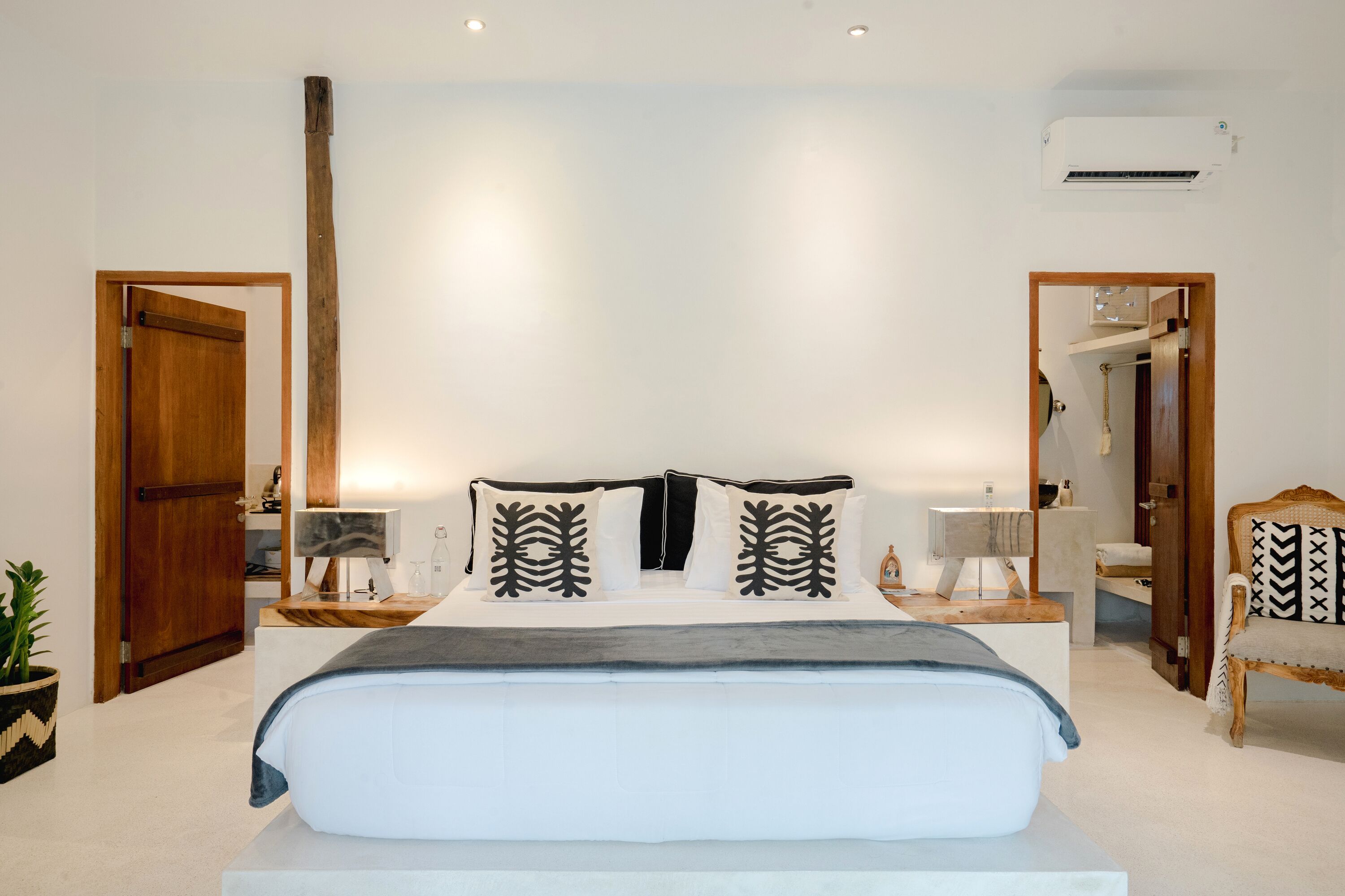 A bedroom with a large bed, bedside tables, a mirror, a wooden chair, and an air conditioning unit.