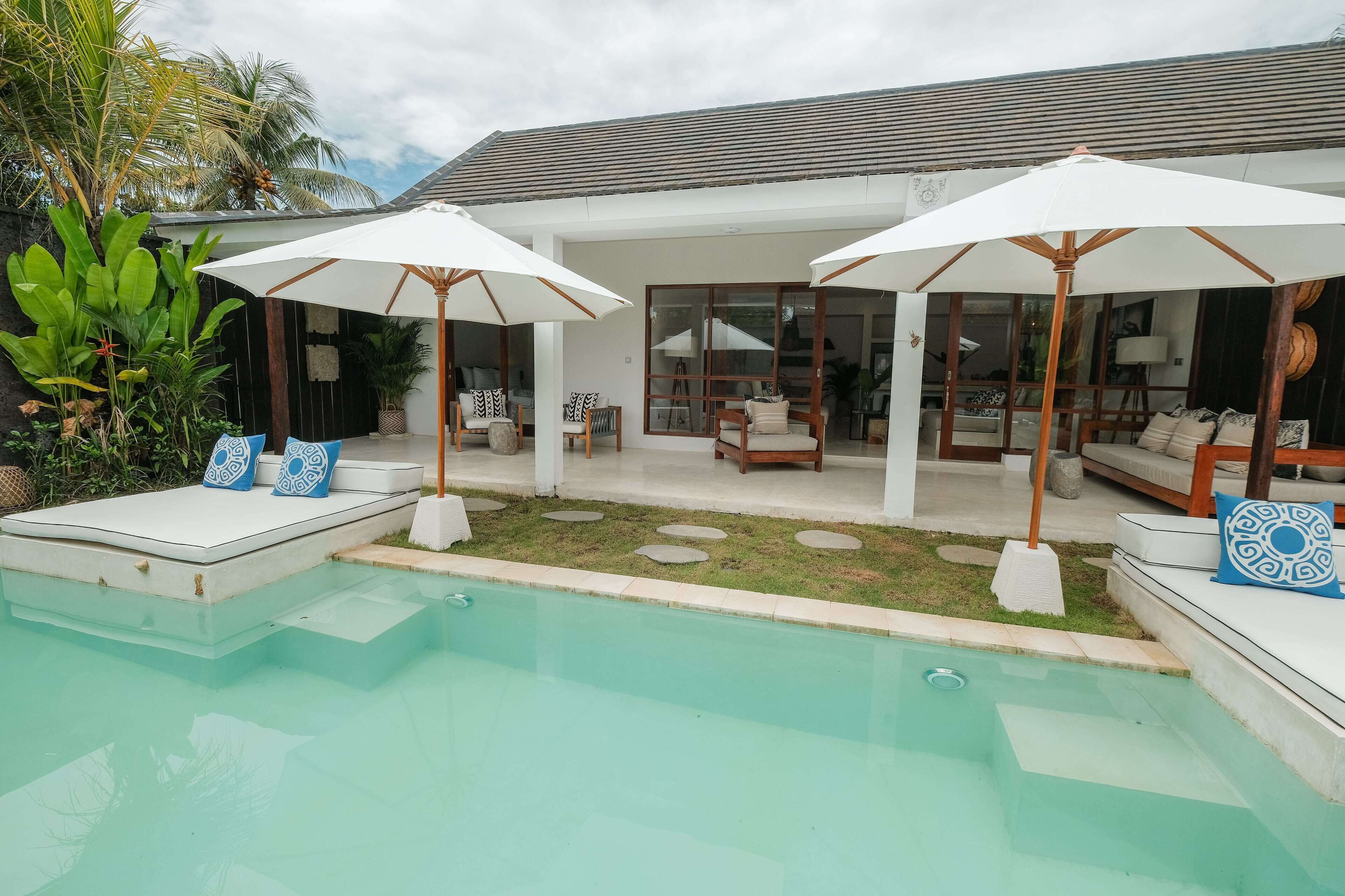 Un área junto a la piscina con sillones de descanso y sombrillas, una zona de estar cubierta con sofás, y vista de la casa al fondo.