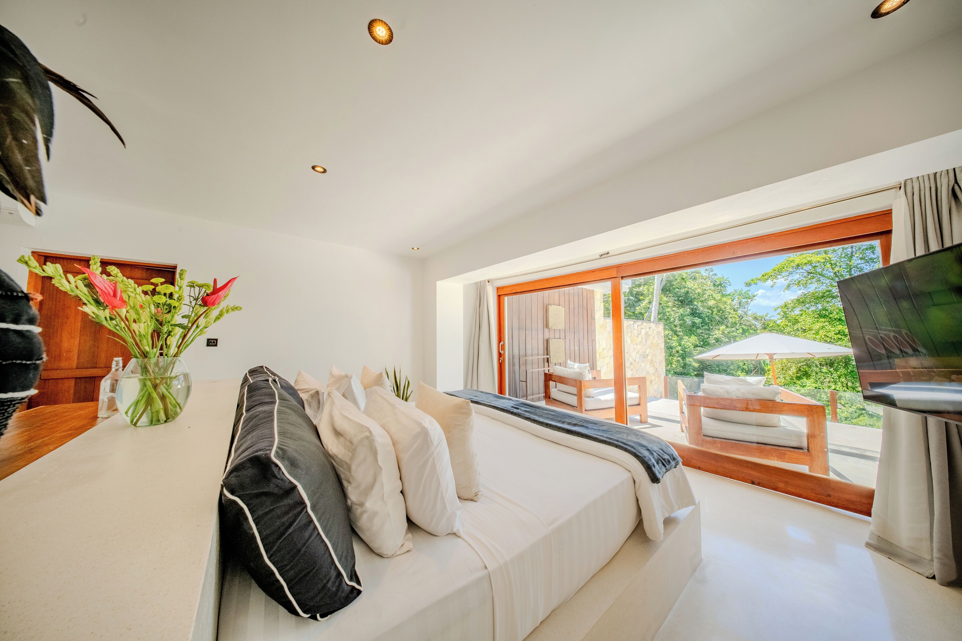 A modern bedroom with a large bed, a TV, a wooden table with a vase of flowers, and a view of the outdoors.