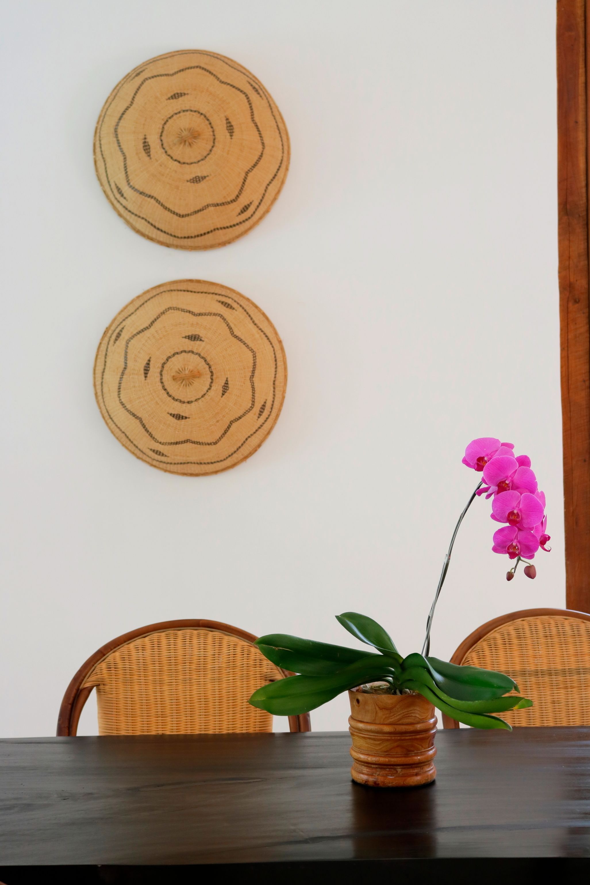 Une table à manger est agrémentée d’un vase en bois contenant une orchidée rose. Deux sous-verres en bois sont placés sur le mur, au-dessus de la table.